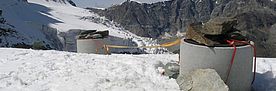 Auf einem schneebedeckten Hang am Matterhorn sind zwei Betonrohre senkrecht in den Schnee und den darunterliegenden Permafrost gegraben. Ihre Metalldeckel sind mit grossen Steinen beschwert, die beiden Rohre sind mit einer losen Leine verbunden. Im Hintergrund der Bohrlöcher erstreckt sich ein Gletscherabbruch und dahinter eine schroffe Felswand.