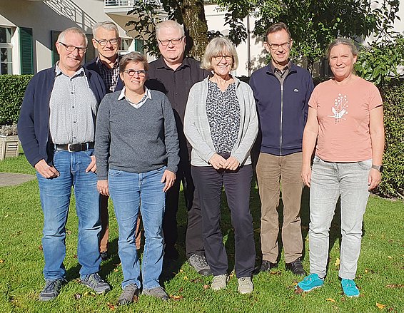 Un groupe de huit personnes se tient debout sur une pelouse verte, souriant. Ils portent des vêtements variés, allant de pulls à des t-shirts, et se tiennent côte à côte, avec un arbre au fond et un bâtiment flou derrière eux.