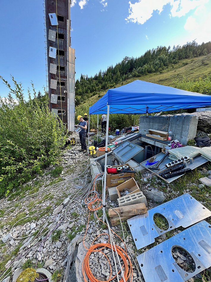 A construction site with a tall, partially completed structure on the left, surrounded by greenery. A blue canopy is set up in the foreground, with various tools and materials like metal sheets and wires scattered around. A person is working nearby.