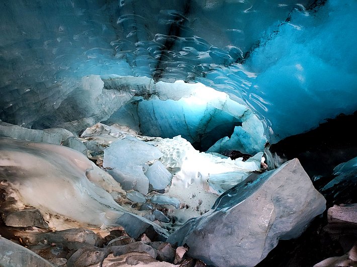 Interior of an ice cave with bluish ice, large ice blocks, and an opening letting in light.
