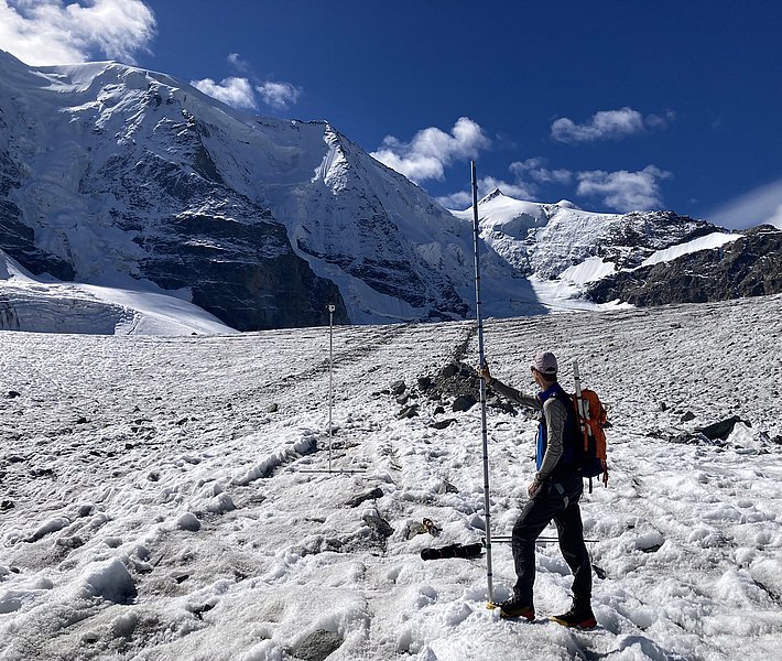 Person with orange backpack stands on snowy glacier holding a long pole, snow-covered mountains visible under blue sky in background.