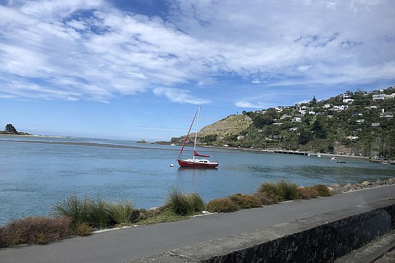 Ein rotes Segelboot schwimmt ruhig im Wasser einer Bucht. Im Hintergrund sind sanfte Hügel zu sehen, auf denen verstreut einige Häuser zu sehen sind . Der Himmel ist teilweise bewölkt.