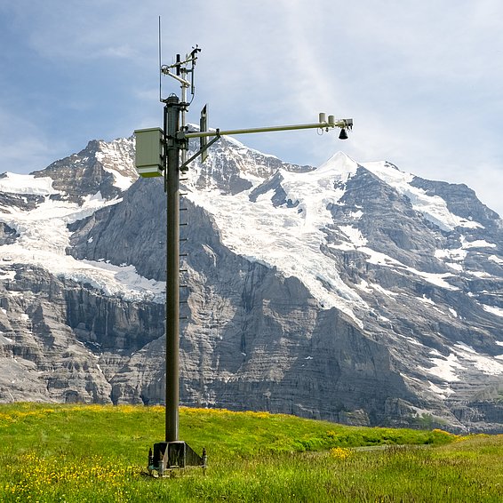 Palo di misurazione con sensori su un prato verde davanti a montagne innevate sotto un cielo blu.