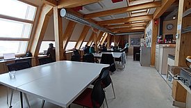 Großer Raum mit Holzbalken, mehreren weißen Tischen und schwarzen Stühlen, Personen arbeiten an Laptops an Fensterplätzen, Küchenbereich mit Geräten und Uhr an der Wand