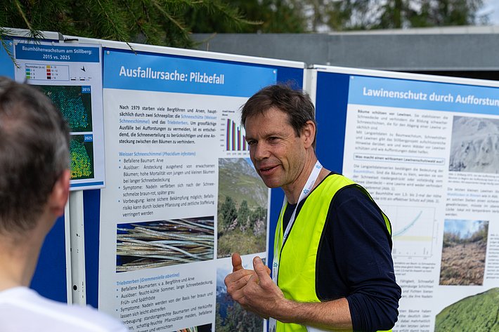 Ein Mann in einer gelben Sicherheitsweste steht vor zwei Informationsständen. Die Stände zeigen Grafiken und Texte über Pilzbefall und Lawinenschutz.
