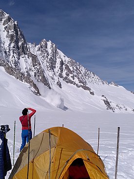 A person in a red shirt and blue pants stands near a yellow tent, gazing towards a snow-covered mountain. In the background, snow is falling from the mountainside, indicating an avalanche. The scene is set in a snowy landscape under a clear sky.