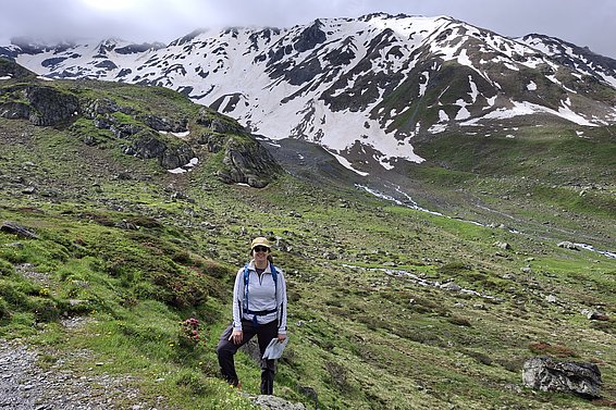 Green mountain landscape with snow on the peaks. Maria standing in the foreground in hiking gear.