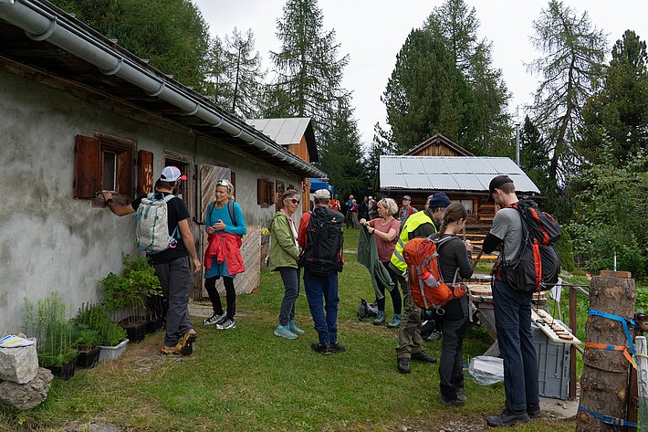 Gruppe von Personen in Outdoor-Bekleidung vor einem alten Stall. Einige stehen, andere sitzen oder lehnen an einem Tisch mit Materialien.