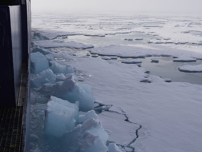 Eisblöcke und gefrorene Wasserflächen am Rand eines Schiffs, mit einer schneebedeckten Landschaft im Hintergrund.