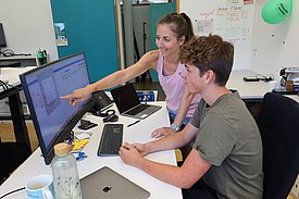 Deux personnes travaillent ensemble à un bureau. Une femme montre quelque chose sur un écran d'ordinateur à un jeune homme. Ils semblent concentrés et échangent des idées, entourés de matériel informatique et de fournitures de bureau.