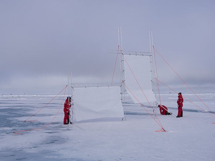 Zwei große, weiße Leinwände auf einem gefrorenen Gewässer, gesichert mit orangefarbenen Seilen. Zwei Personen in roten Anzügen montieren die Leinwände.