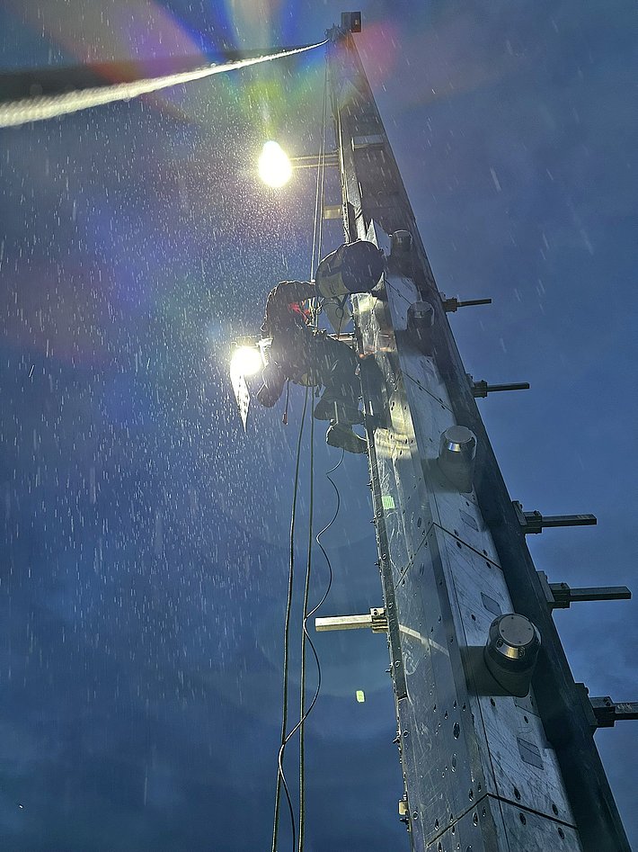 A person is climbing a tall utility pole in rainy weather. The scene is illuminated by overhead lights, creating a dramatic effect with raindrops falling around them against a dark, cloudy background.