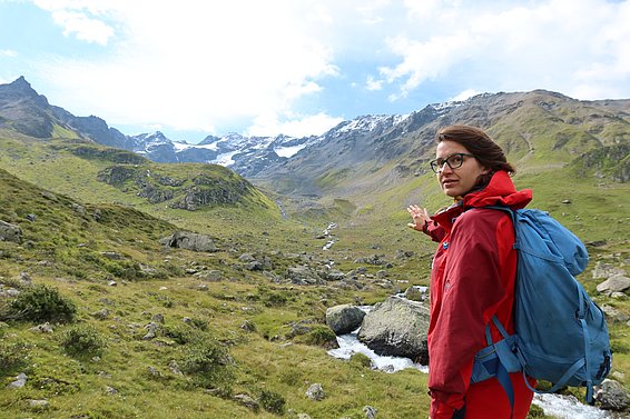 A woman with a red jacket and blue rucksack points to a mountain landscape with green grass, rocks and snow-covered peaks under a blue sky.