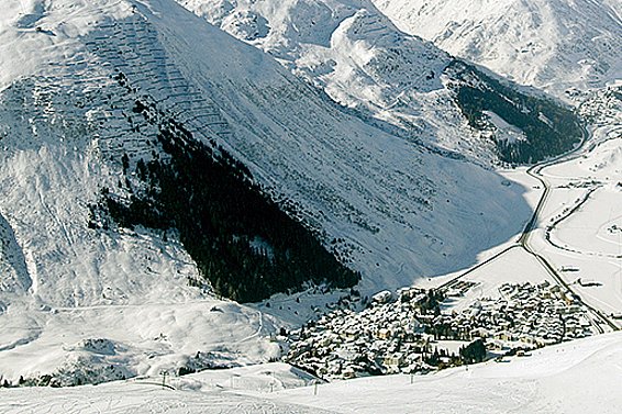 Das Bild zeigt den Blick von oben auf Andermatt  und den darüberliegenden Bannwald. Alle Berghänge sind schneebedeckt, nur direkt über dem Dorf kontrastiert der dunkle Schutzwald mit dem weissen Hintergrund.