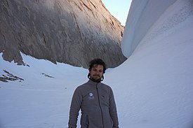 Mann in grauer Polar Exploration Jacke steht vor schneebedecktem Fels und großer Schneeformation in Gebirgslandschaft