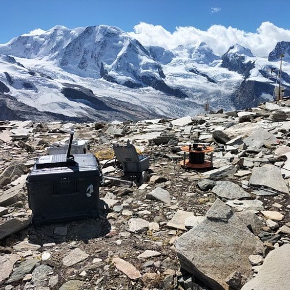 Apparecchio con antenna, sedia e avvolgicavo su terreno roccioso davanti a montagne innevate sotto un cielo blu.