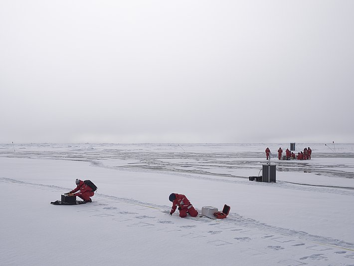 Zwei Personen in roten Anzügen arbeiten auf einer schneebedeckten Fläche. Eine Person sitzt, die andere ist in gebückter Haltung. Im Hintergrund sind mehrere weitere Personen zu sehen.