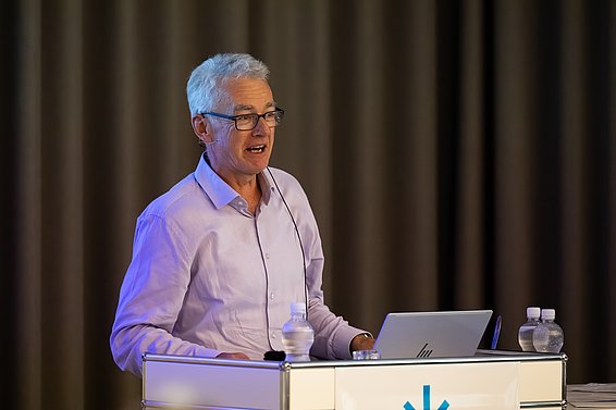 Älterer Mann mit grauen Haaren und Brille steht hinter einem Rednerpult mit Laptop und Wasserflaschen, trägt ein hellviolettes Hemd.