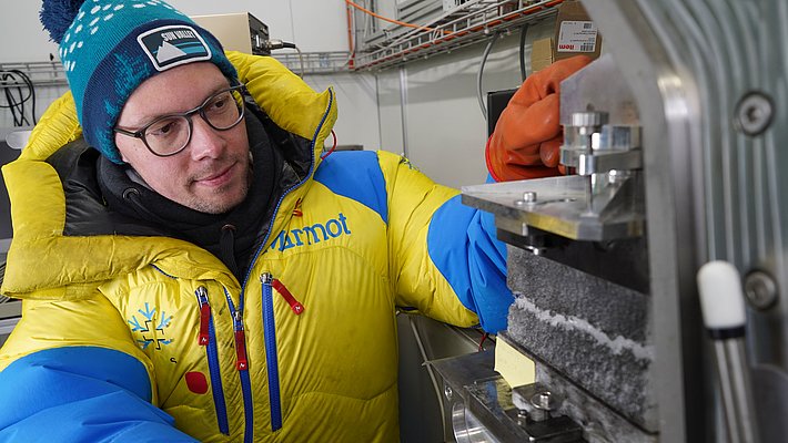 Ein Wissenschaftler in einer bunten Winterjacke arbeitet an einem Gerät, das eine Schneeprobe drückt. Er trägt Handschuhe und eine Mütze, während er konzentriert die Maschine bedient. Der Hintergrund zeigt eine Laborausrüstung.