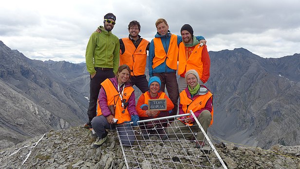 Sieben Personen, sechs davon in einer orangen Warnweste, stehen auf einem Berggipfel. Sie halten einen grossen Metallrahmen, der mit Schnüren in kleine Quadrate unterteilt ist. Mithilfe dieses Gitters werden Vegetationsaufnahmen gemacht.