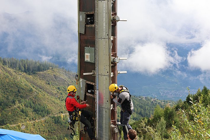 Two climbers in helmets are ascending a tall, vertical structure on a mountainous landscape. One climber is positioned higher up on the structure, while the other climbs below. The background features green hills and a cloudy sky.