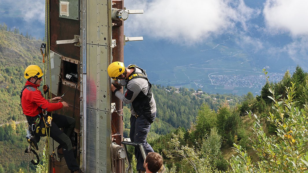 Two climbers in helmets are ascending a tall, vertical structure on a mountainous landscape. One climber is positioned higher up on the structure, while the other climbs below. The background features green hills and a cloudy sky.