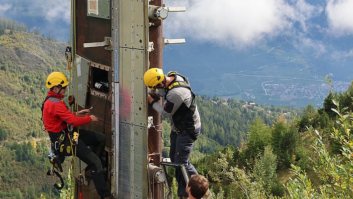 Zwei Personen in Kletterausrüstung besteigen einen hohen, vertikalen Mast in einer bergigen Landschaft. Die Umgebung ist von Bäumen und Wolken umgeben, während die Kletterer die Struktur erklimmen.