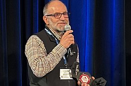 Ein Mann in Jeans, kariertem Hemd und schwarzer Weste mit dem Logo des SLF hält einen Preis in der Hand und spricht in ein Mikrophon.