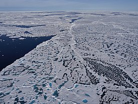 Luftaufnahme einer arktischen Landschaft mit schneebedecktem Eis, Wasserflächen und verschiedenen Eismustern. Der Himmel ist klar und blau.