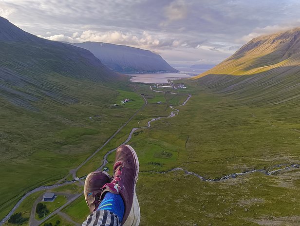 A pair of feet in colorful socks rests on a rocky ledge, overlooking a vast valley. The landscape features green hills, a winding river, and a small settlement nestled in the distance, under a partly cloudy sky.