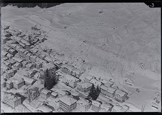Black-and-white aerial view of an avalanche cone on a mountainside next to a village with buildings and roads