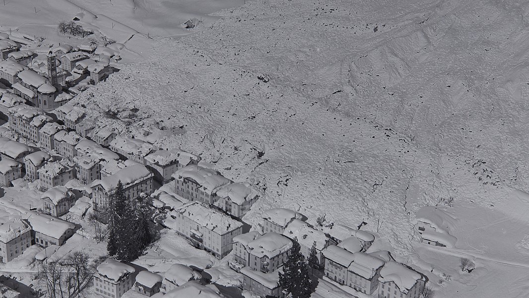 Schwarz-weiße Luftaufnahme eines Lawinenkegels an einem Berghang neben einem Dorf mit Gebäuden und Straßen