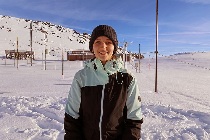 A young person stands smiling in a snowy landscape, wearing a black and turquoise jacket and a black beanie. In the background, snow-covered hills and equipment can be seen, indicating a winter sports setting. Skis and poles are partially visible, emphasizing the outdoor activity theme.