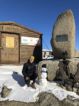 Eine Frau kniet vor einem Haus neben einem Schneemann am Boden und lacht in die Kamera. Daneben steht ein grosser Stein mit einer Inschrifttafel.