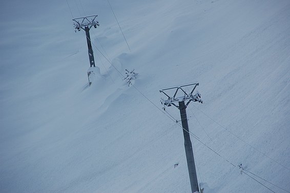 Zwei Masten stehen in einer schneebedeckten Landschaft. Die Masten sind dunkel und tragen jeweils mehrere Querstreben, an denen Seile befestigt sind. Die Seile verlaufen diagonal durch das Bild. Der Hintergrund ist überwiegend weiß und zeigt eine glatte Schneefläche.