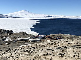 The Mario Zucchelli Station in Tera Nova Bay, surrounded by rocky terrain, with a partially frozen sea and a snow-covered mountain in the background.