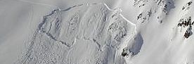 Das Bild zeigt einen schneebedeckten Hang, in dessen oberem Viertel eine Schneebrettlawine angerissen ist.  Eine Aufstiegsspur durchquert die Gleitfläche und im unberührten Schnee links von der Lawine schlängelt sich eine Abfahrtsspur den Hang hinunter.