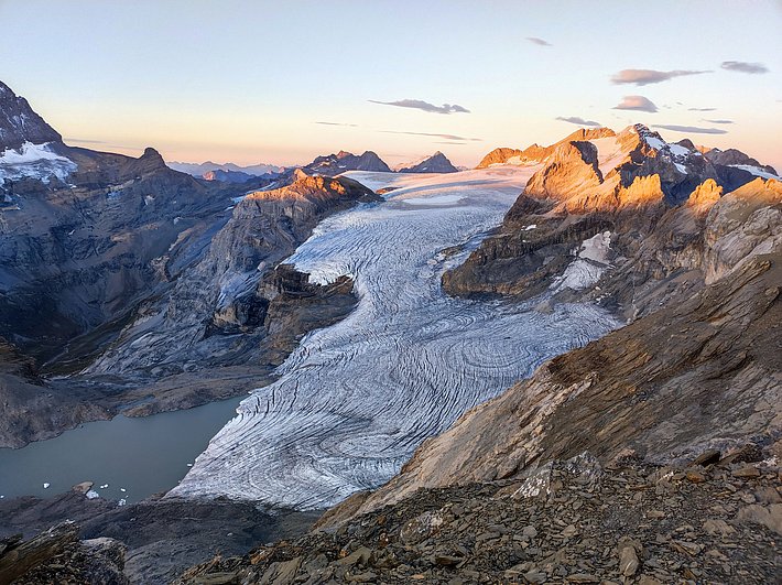 Glacier flowing between rocky mountains at sunset with orange light on peaks and a small lake on the left.
