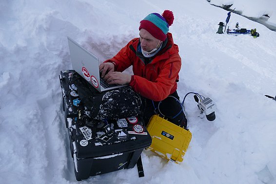 Una persona in abbigliamento invernale scrive su un laptop, seduta sopra una cassa nera in mezzo alla neve. Sullo sfondo si vedono attrezzature da campo. L'ambiente è innevato e sembra essere una zona remota.