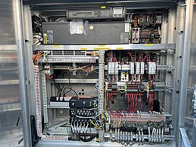 An open electrical control cabinet displaying various components, including circuit boards, wiring, and relay modules. The interior is organized with neatly arranged cables and connectors, showcasing a complex network of electrical connections for operational functions.