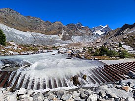 Berglandschaft mit schneebedeckten Gipfeln, einem Fluss mit Wehr und felsigem Ufer unter klarem blauem Himmel