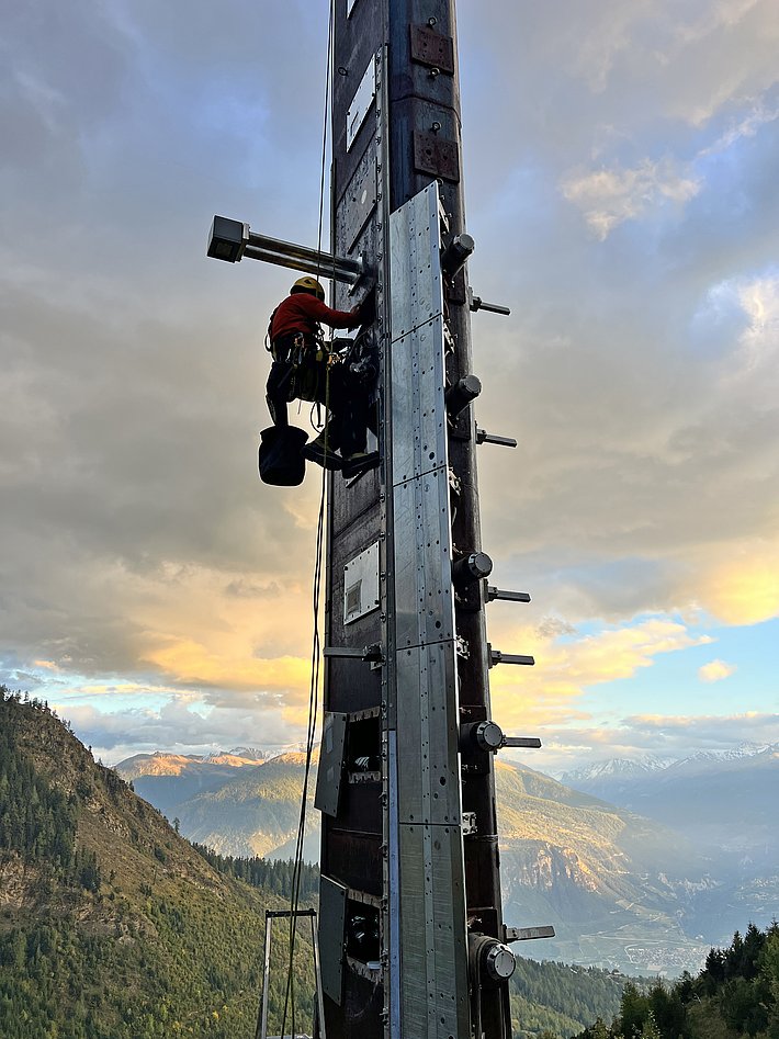 A climber in safety gear scales a tall structure on a mountain, with scenic views of valleys and mountains in the background. The sky is partly cloudy, casting a warm glow over the landscape.
