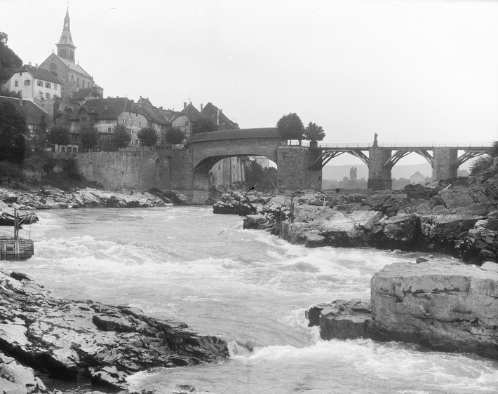 Der Rhein mit Felsen und schnell fliessendem Wasser ist im Vordergrund, im Hintergrund sind eine Brücke und Laufenburg zu sehen.