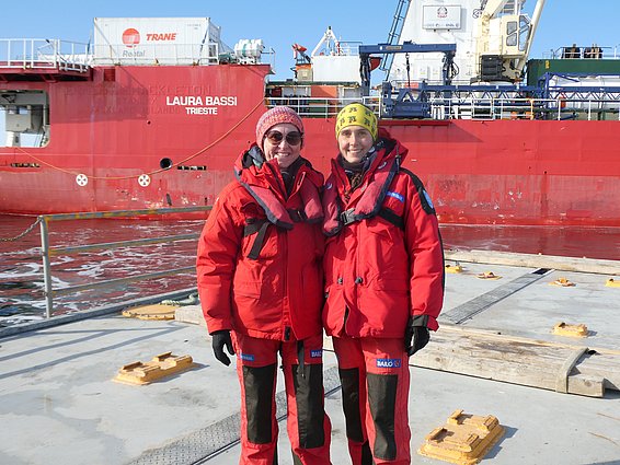 Zwei Frauen in roten Anzügen lachen in die Kamera. Im Hintergrund ist ein ebenfalls rote Schiff zu sehen, dass den Namen Laura Bassi auf dem Bug trägt.