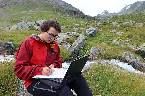 A woman in a red outdoor jacket sits on rocks in a mountainous landscape, writing in a notebook, with a laptop on her lap.