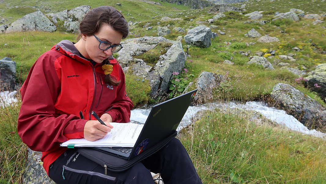 Frau in roter Outdoorjacke sitzt auf Felsen in bergiger Landschaft, schreibt in Notizbuch, Laptop auf Schoß.