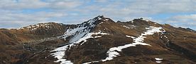 Das Bild zeigt ein alpines Skigebiet in herbstlichen Farben. Nur rund um die Schneelanzen ist der Boden bereits weiss und lässt den Verlauf der Pistenlinie erahnen.