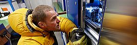 Im Kältelabor kniet ein Forscher im dicken gelben Daunenanzug vor einem offenen Computertomographen und bereitet sich darauf vor, eine Schneeprobe im Gerät zu installieren.