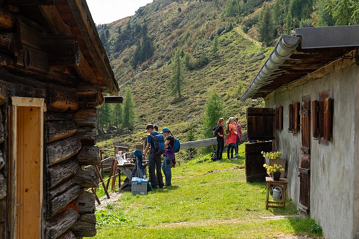 Zwei Gebäude aus Holz und Stein stehen auf einer Wiese. Eine Personengruppe steht im Freien. Die Landschaft ist hügelig und bewaldet.