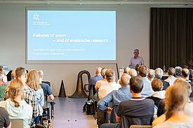 Speaker stands in front of screen with presentation on the topic “Failures of snow – and of avalanche research” in front of an audience in a conference room.